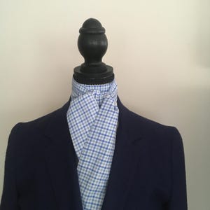 May include: A navy blue blazer with a light blue and white checkered ascot. The ascot is draped around a black mannequin head. The blazer has a classic cut and is made of a smooth fabric.