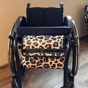 Może przedstawiać: Czarny wózek inwalidzki z czarnym siedziskiem i oparciem. Z przodu przymocowana jest torba z nadrukiem w cętki. Torba ma czarny pasek i srebrne zapięcie. Koła są srebrne i czarne.