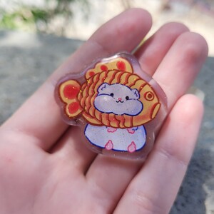 May include: A clear resin charm featuring a cartoon hamster wearing a golden fish-shaped hat with red and orange accents. The hamster is white with pink cheeks and paws. The charm has a glittery finish.