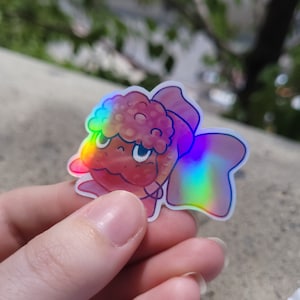 May include: A pink and brown cartoon sticker of a creature with a bow. The sticker has a holographic finish.