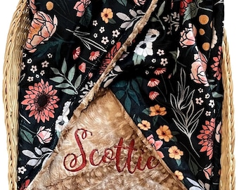 May include: A black and brown baby blanket with a floral pattern and the name "Scottie" embroidered in red on the brown side. The blanket is folded and placed in a wicker basket.