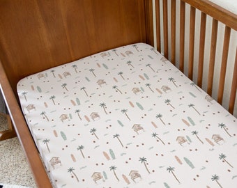 May include: A white crib sheet with a pattern of palm trees, surfboards, and small houses. The sheet is in a wooden crib with a brown wooden frame.
