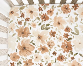 May include: A white fitted crib sheet with a watercolor floral pattern in shades of brown and beige. The sheet features a variety of flower shapes and sizes, with green leaves scattered throughout.