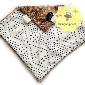 May include: A white and brown baby blanket with a black and white geometric pattern. The blanket has a brown faux fur trim and a pacifier attached to the corner. The tag says "NEW! Design Update" and "no snaps".