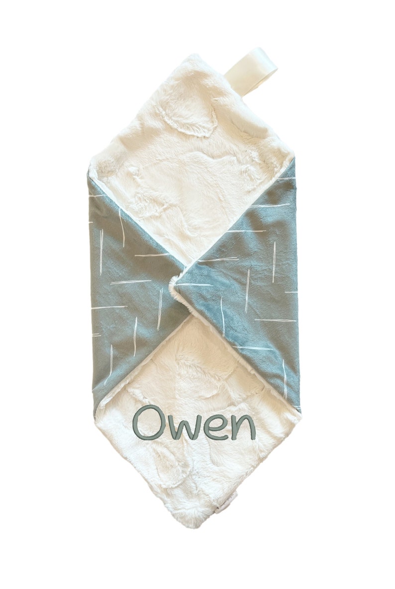Puede incluir: Una manta suave para beb&eacute;, blanca y gris con un lazo de sat&eacute;n blanco. La manta tiene un patr&oacute;n geom&eacute;trico en un lado y una textura suave y peluda en el otro lado. La manta est&aacute; personalizada con el nombre "Owen".
