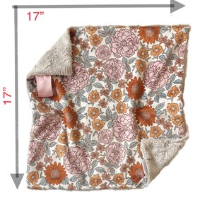 May include: A square blanket measuring 17 inches by 17 inches, featuring a floral pattern with pink and orange flowers and light blue leaves on a cream background. The blanket has a soft, textured beige underside and a small pink fabric tag.