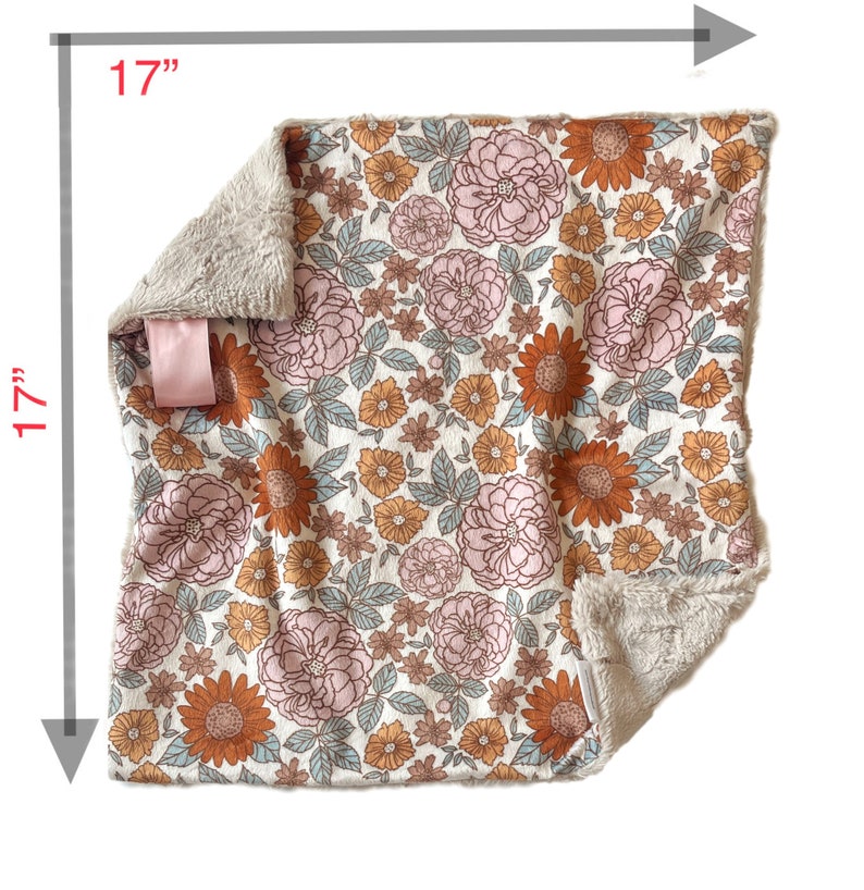 May include: A square blanket measuring 17 inches by 17 inches, featuring a floral pattern with pink and orange flowers and light blue leaves on a cream background. The blanket has a soft, textured beige underside and a small pink fabric tag.