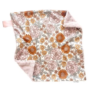May include: A square security blanket featuring a floral pattern with rust-orange sunflowers, light pink roses, and sage green leaves on a cream background. The reverse side is a soft, textured light pink plush fabric with a small pink ribbon tag.