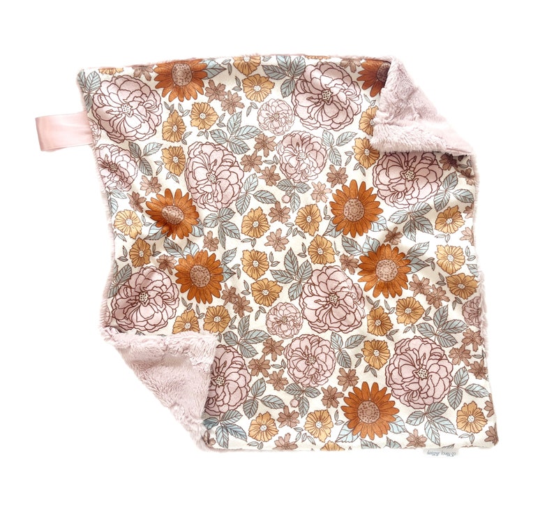 May include: A square security blanket featuring a floral pattern with rust-orange sunflowers, light pink roses, and sage green leaves on a cream background. The reverse side is a soft, textured light pink plush fabric with a small pink ribbon tag.
