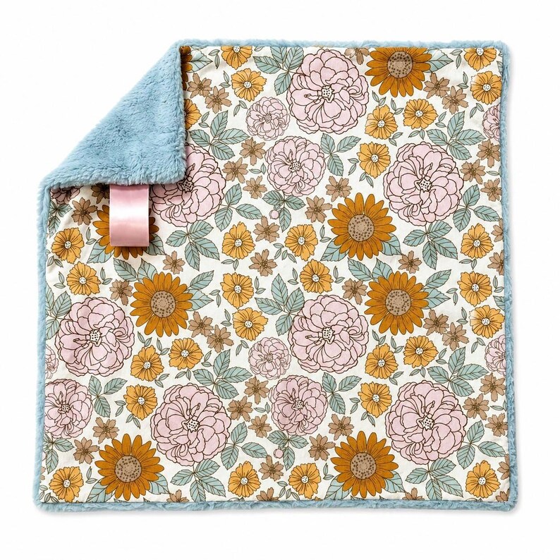 May include: A square baby blanket with a floral pattern on a white background featuring pink roses, mustard yellow sunflowers, and sage green leaves. The reverse side is a solid light blue faux fur fabric with a pink satin ribbon tag attached.