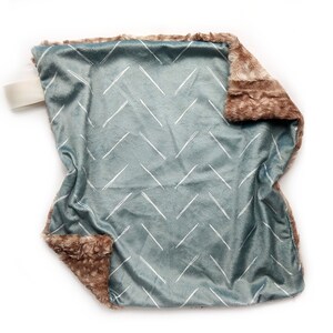 Puede incluir: Una manta suave para beb&eacute;, de color azul y marr&oacute;n, con un patr&oacute;n de chevron blanco. La manta est&aacute; hecha de un tejido de felpa y tiene un borde peludo.