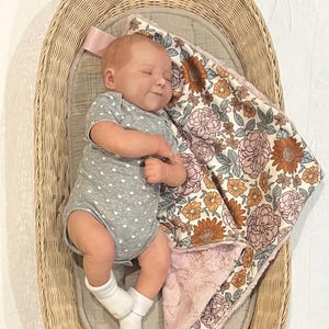 May include: A realistic baby doll with light skin and short hair, wearing a gray short-sleeved bodysuit with a white heart pattern and white socks, lying in an oval wicker basket next to a soft blanket featuring a floral print in orange and pink.
