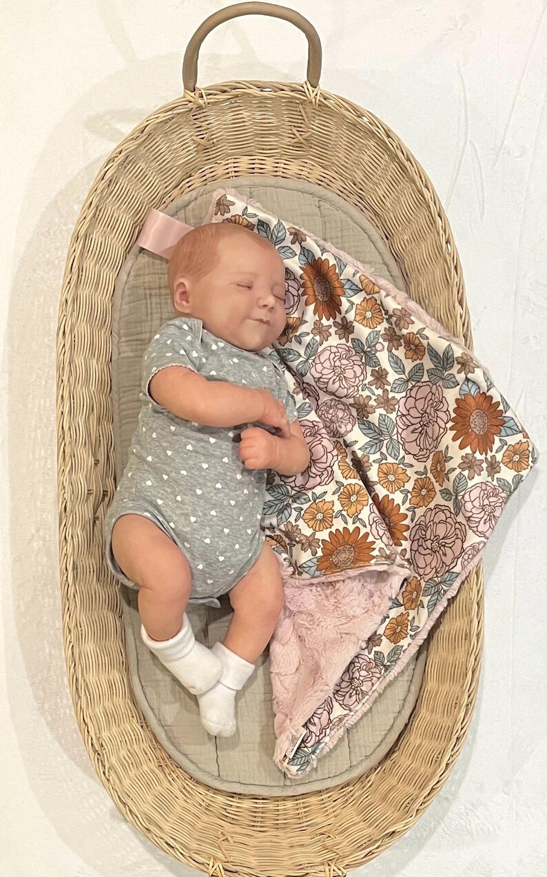 May include: A realistic baby doll with light skin and short hair, wearing a gray short-sleeved bodysuit with a white heart pattern and white socks, lying in an oval wicker basket next to a soft blanket featuring a floral print in orange and pink.