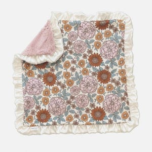 May include: A square baby blanket featuring a floral pattern with pink, rust-orange, and muted blue flowers on a cream background. The blanket has a ruffled cream satin border and a soft, textured pink faux fur lining visible on the folded corner.