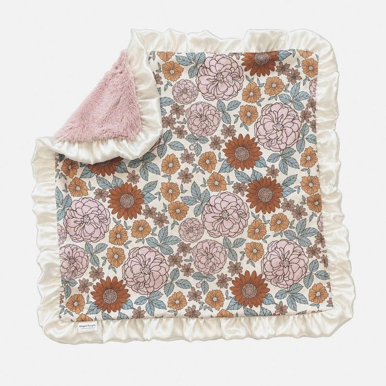 May include: A square baby blanket featuring a floral pattern with pink, rust-orange, and muted blue flowers on a cream background. The blanket has a ruffled cream satin border and a soft, textured pink faux fur lining visible on the folded corner.