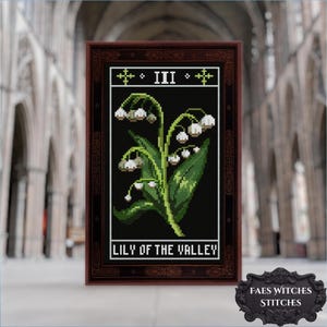 May include: Cross-stitch art of Lily of the Valley flowers in a dark wooden frame. The design features white bell-shaped flowers, green leaves, and the text "LILY OF THE VALLEY". The artwork is framed with a decorative border and the number "III" at the top.
