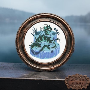 Pode incluir: Uma peça de ponto cruz emoldurada com um dragão em tons de verde-azulado e verde, sobre um fundo azul claro. O dragão está rodeado por água e vegetação. A moldura redonda tem um acabamento em bronze escuro. O texto "FAES WITCHES STITCHES" está na parte inferior.