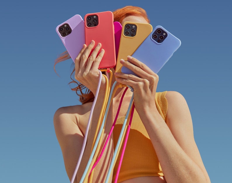 K&ouml;nnte beinhalten: Eine Person h&auml;lt vier bunte Handyh&uuml;llen in ihren H&auml;nden. Die H&uuml;llen sind rosa, gelb, blau und lila. Die H&uuml;llen sind an bunten Handyriemen befestigt.
