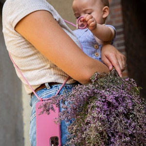 K&ouml;nnte beinhalten: Eine Frau tr&auml;gt eine rosa Handyh&uuml;lle mit einem Band um den Hals. Die Handyh&uuml;lle ist an einem rosa Band befestigt, das um ihren Hals geht. Sie h&auml;lt ein Baby in ihren Armen und einen Strau&szlig; lila Blumen.