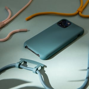 K&ouml;nnte beinhalten: Eine tealblaue Handyh&uuml;lle mit einem Ausschnitt f&uuml;r das Kameraobjektiv. Die H&uuml;lle liegt auf einer hellblauen Oberfl&auml;che mit einem blauen und wei&szlig;en Lanyard daneben.