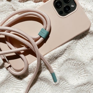 K&ouml;nnte beinhalten: Eine hellrosa Handyh&uuml;lle mit einem schwarzen Kameraobjektiv und einem hellrosa Handyband mit t&uuml;rkisfarbenen Akzenten. Auf der Handyh&uuml;lle steht "PopSocket".