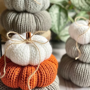 May include: A stack of three knitted pumpkins in white, orange, and gray, featuring a cinnamon stick stem and a tied jute twine bow. The pumpkins have a ribbed texture and are arranged in a vertical pile, with another similar stack visible nearby.