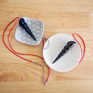May include: Two black spiral shell pendants on white and blue patterned dishes. One pendant is on a red cord with a silver chain, the other is on a red cord.