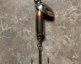 May include: A silver fishing lure with a bullet-shaped body and a silver blade. The lure has a triple hook.