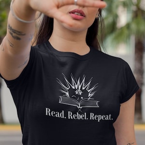 Könnte beinhalten: Ein schwarzes T-Shirt mit einer weißen Grafik eines Buches mit einem Stern und dem Text "Read. Rebel. Repeat."