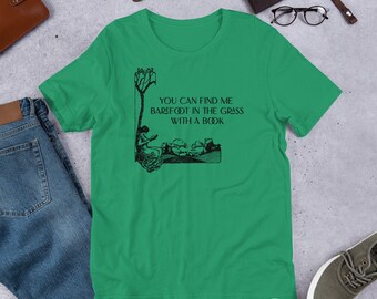 May include: A green t-shirt featuring a black illustration of a person sitting in the grass reading a book. The text on the shirt reads "You can find me barefoot in the grass with a book."