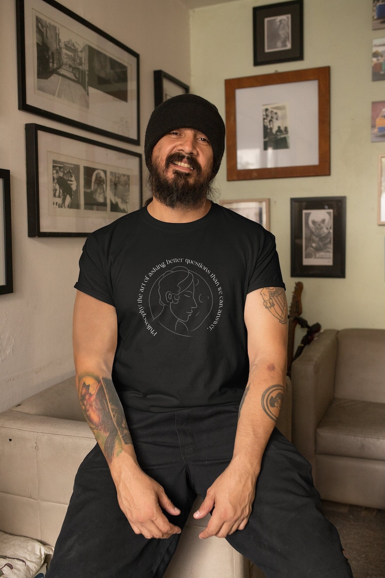 May include: A person wearing a black t-shirt with a white graphic design that says "Philosophically, the art of asking better questions than we can answer." The graphic design features a line drawing of a person's head with a moon and stars.