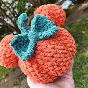 May include: A crocheted pumpkin-shaped plush toy with a green bow. The toy is orange and has a textured surface.