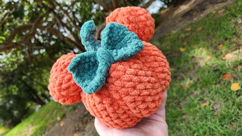 May include: A crocheted pumpkin-shaped plush toy with a green bow. The toy is orange and has a textured surface.