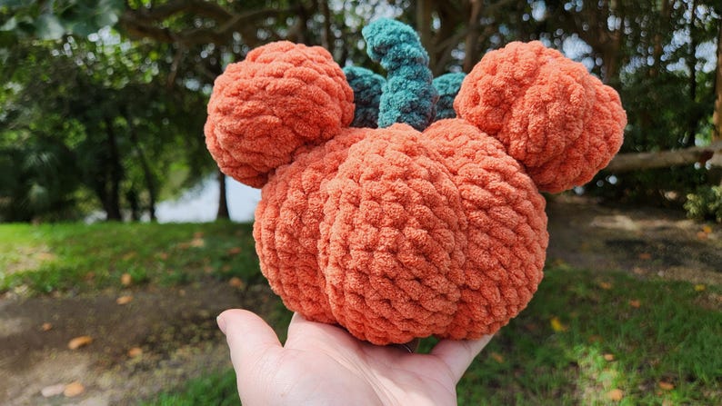 May include: A crocheted pumpkin with a green stem and orange body. The pumpkin is shaped like a Mickey Mouse head.