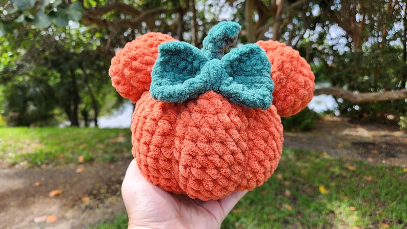 May include: A crocheted pumpkin with a green bow, resembling a popular cartoon character. The pumpkin is orange and the bow is a dark green.