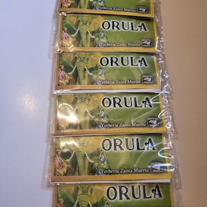 Può includere: Una pila di confezioni sigillate con la parola "ORULA" a caratteri grandi. Ogni confezione presenta un'illustrazione di un elefante e figure, con il testo "Yerberia Zanta Muerte" e "Hecho en Mexico."