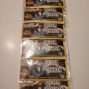 May include: Six packets of "QUITA VICIOS" are stacked, each with a picture of glasses with a brown liquid and the text "Yerberia Zanta Muerte". The packets are sealed in clear plastic. The text is in a stylized font.
