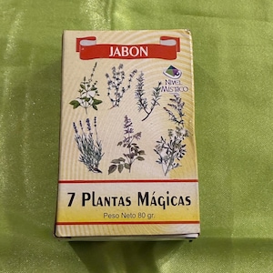 Op de afbeelding: Een rechthoekige zeepdoos met het woord "JABON" in het rood. De doos toont illustraties van zeven planten en de tekst "7 Plantas Mágicas". Het nettogewicht is 80 g.