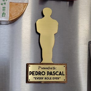 Könnte beinhalten: Eine goldfarbene Trophäe in Form einer Person, montiert auf einem Holzsockel. Auf dem Sockel steht "Presented to: PEDRO PASCAL" und "EVERY ROLE EVER" in weißer Schrift. Die Trophäe ist auf einer metallischen Oberfläche befestigt.