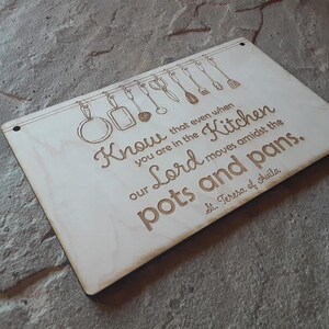 May include: A light-colored wooden sign with engraved kitchen utensils hanging above a quote. The quote reads "Know that even when you are in the kitchen our Lord moves amidst the pots and pans." The sign is attributed to St. Teresa of Avila.