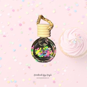 May include: A clear glass car air freshener with colorful confetti inside, a wooden top, and a braided rope. A cupcake with pink frosting and sprinkles is next to the air freshener. The background is pink with confetti and the text "Handmade By Hayli".