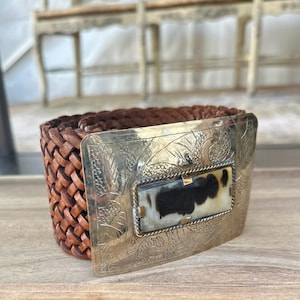 May include: A brown leather belt with a woven design. The belt features a large, ornate silver buckle with a rectangular inset of black and white patterned material. The buckle has a floral design.
