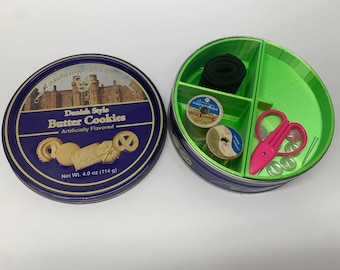May include: A green round tin with three compartments. The compartments contain a roll of black elastic, two spools of thread, a pair of pink scissors, and a few buttons.
