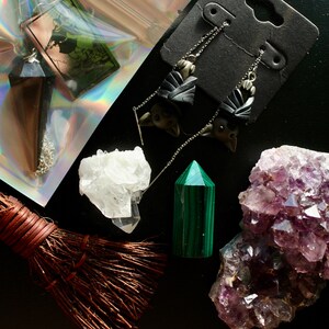 Pode incluir: Uma variedade de itens místicos em uma superfície preta. Inclui um pêndulo, brincos de morcego, um cristal de quartzo, uma torre de malaquita, um aglomerado de ametista roxa e uma pequena vassoura.