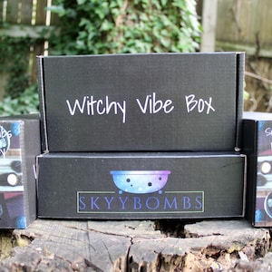 May include: Three black boxes stacked on top of each other. The top box says "Witchy Vibe Box" in white text. The middle box says "SKYBOMBS" in white text with a blue and purple galaxy design. The bottom box says "Support Small Businesses" in white text with a galaxy design.
