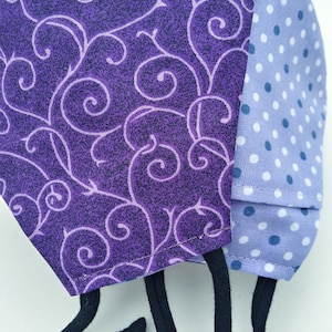 May include: A purple and blue fabric face mask with a swirl pattern on one side and white polka dots on the other side. The mask has black adjustable straps.