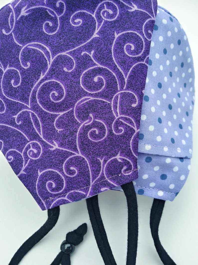 May include: A purple and blue fabric face mask with a swirl pattern on one side and white polka dots on the other side. The mask has black adjustable straps.