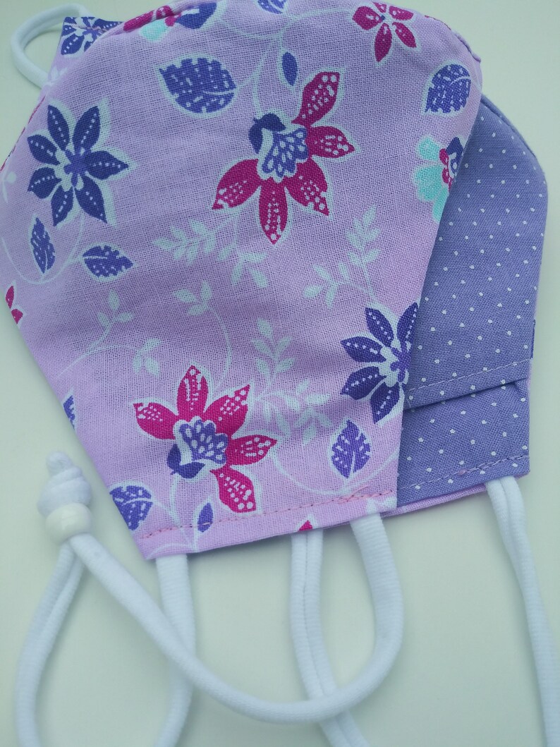 May include: A purple and white floral fabric face mask with a purple polka dot fabric lining. The mask has white elastic ear loops.