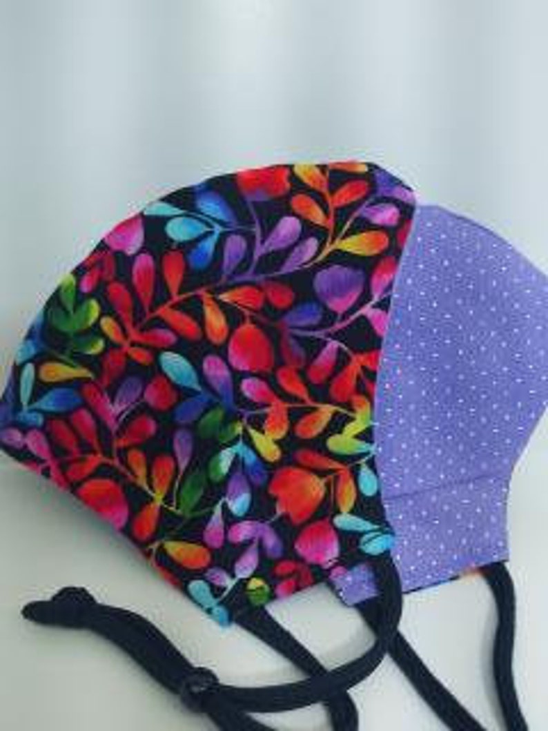 May include: A reusable face mask with a black background and a colorful floral pattern on one side and a purple polka dot pattern on the other side. The mask has black elastic ear loops.