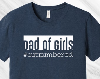 May include: Navy blue t-shirt with the text "dad of girls" in a white rectangular box, and the hashtag "#outnumbered" below. The shirt is made of a soft material.